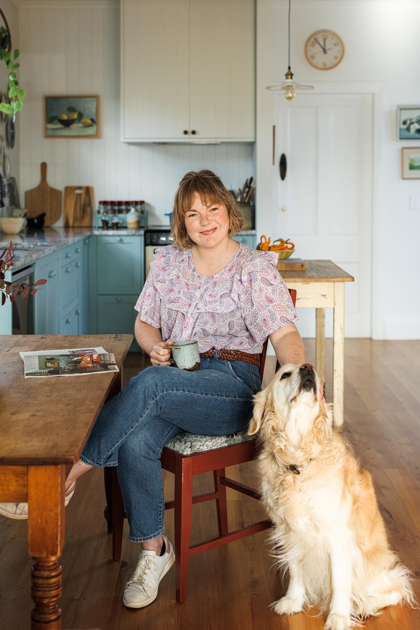 Woman sitting at a kitchen table with a dog, holding a mug.