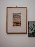 A framed landscape painting with a gold frame and cream mat, hanging on a light gray wall next to another framed artwork.