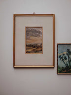 A framed landscape painting with a gold frame and cream mat, hanging on a light gray wall next to another framed artwork.