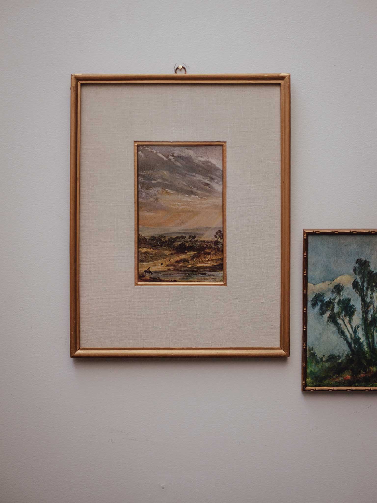 A framed landscape painting with a gold frame and cream mat, hanging on a light gray wall next to another framed artwork.