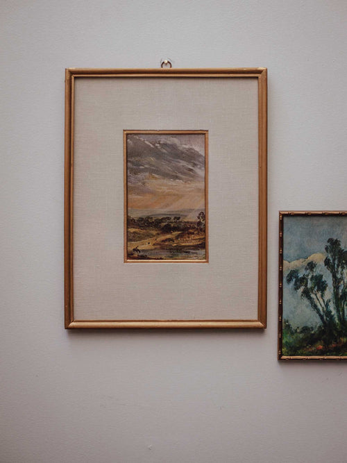 A framed landscape painting with a gold frame and cream mat, hanging on a light gray wall next to another framed artwork.