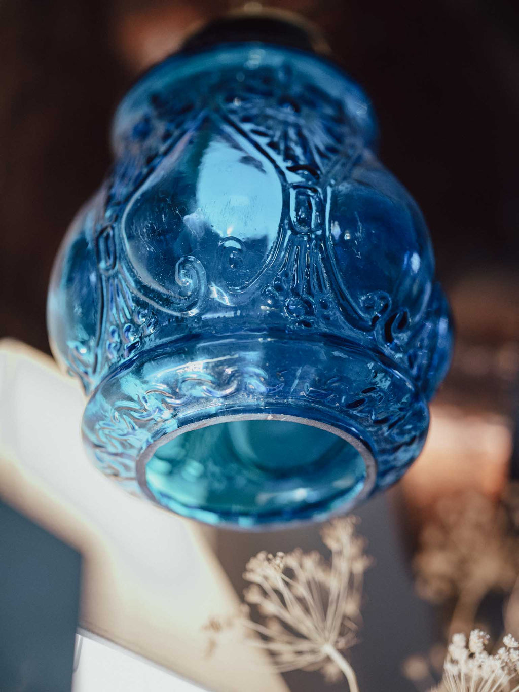 A close-up of an ornate, vintage-style blue glass lamp shade with intricate embossed floral and scrollwork details, set against a dark, blurred background with dried flowers in the foreground.