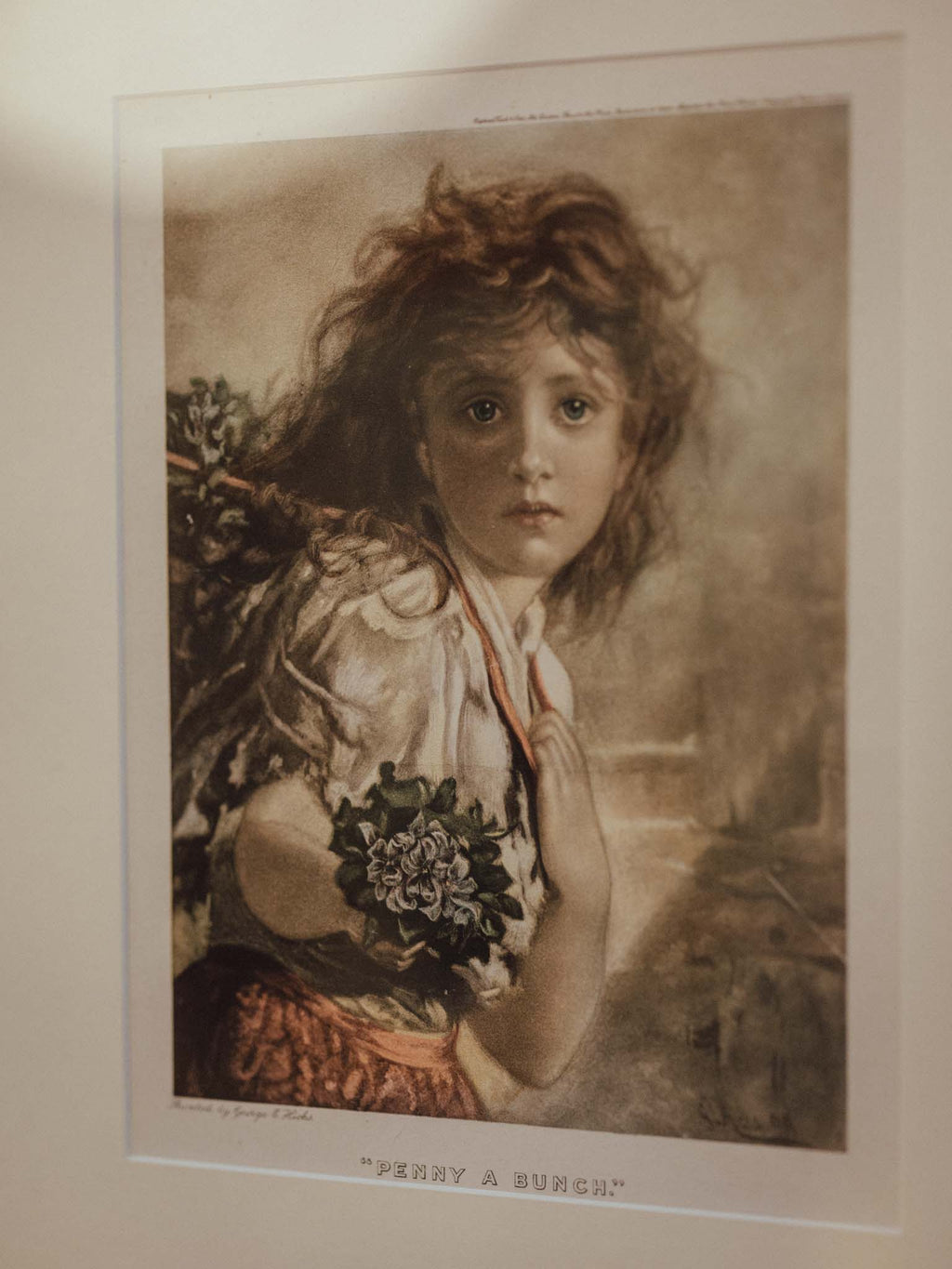 A vintage-style art print featuring a young girl with curly hair, wearing a white blouse and red skirt, holding a bouquet of flowers. The print is mounted on a light-colored wall with a clear protective sleeve.