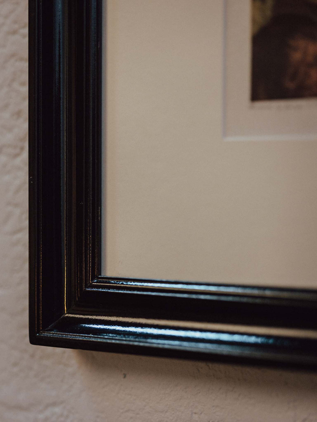 Close-up of a glossy black picture frame corner mounted on a textured beige wall, with a cream mat and a partially visible portrait print.
