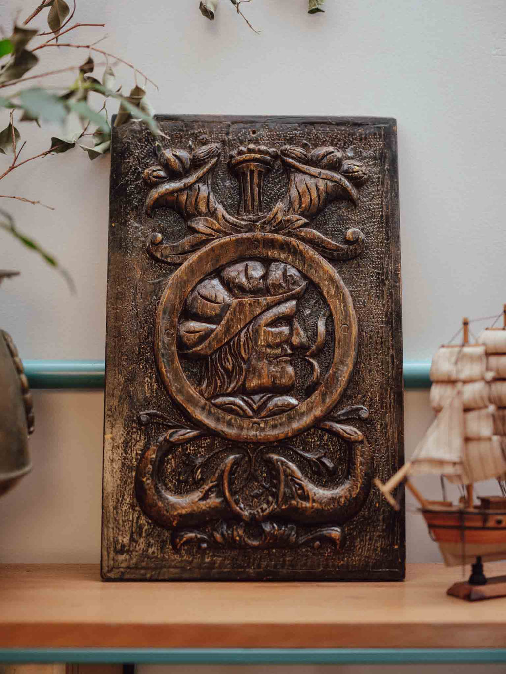A rectangular dark brown carved wooden wall plaque featuring a circular emblem with a figure in profile and ornate scrollwork, displayed on a wooden shelf against a light wall with decorative plants and a model ship nearby.