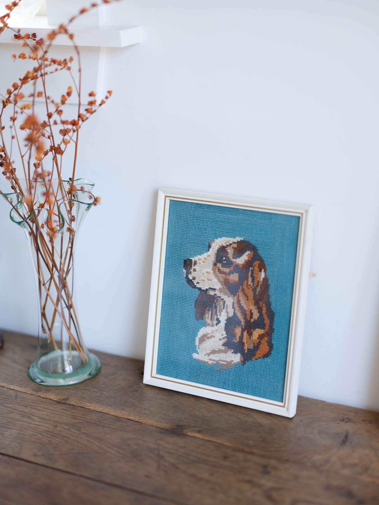 Framed needlepoint of a dog on a wooden surface with a vase of dried flowers.