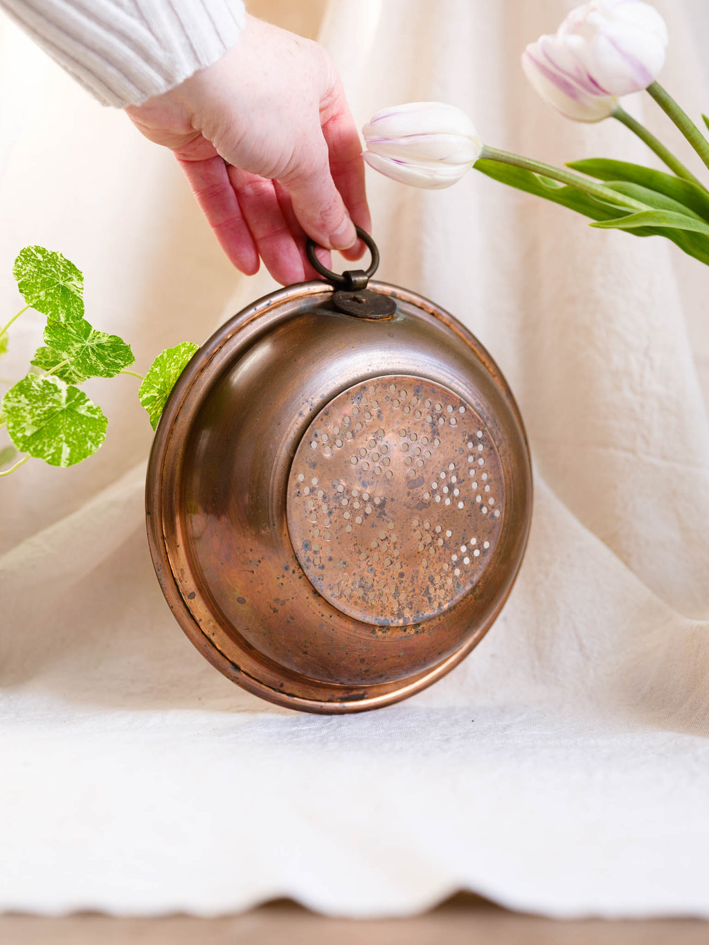 Copper pot held by a hand with tulips and green leaves in the background
