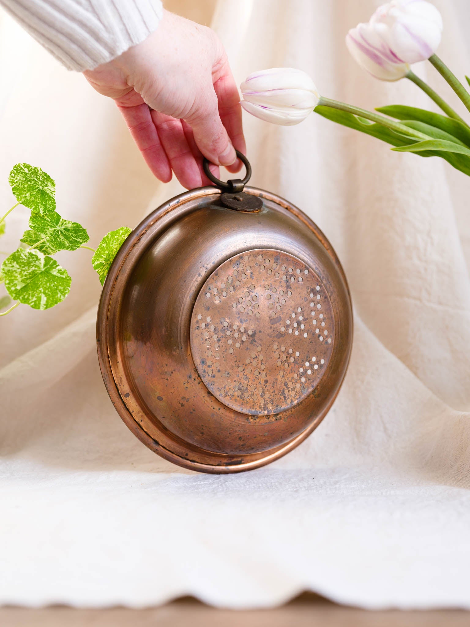 Copper pot held by a hand with tulips and green leaves in the background
