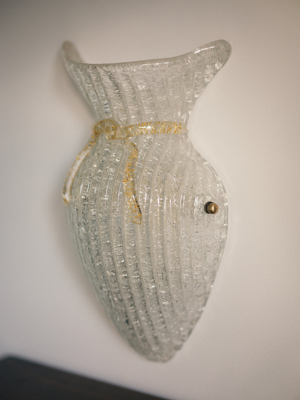 Vintage Murano glass wall sconce with ribbed clear glass and gold ribbon detailing. Italian mid-century wall light, hand-blown using the Graniglia technique. Mounted on a light grey backplate with brass fittings, perfect for hallways, bedrooms, or statement lighting.
