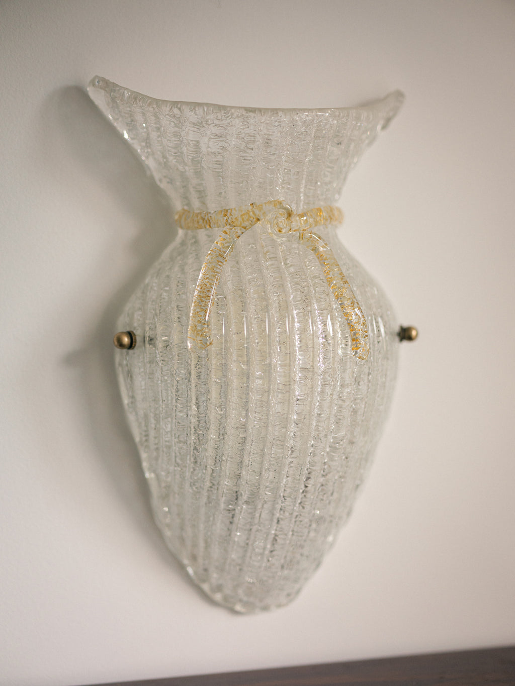 Vintage Murano glass wall sconce with ribbed clear glass and gold ribbon detailing. Italian mid-century wall light, hand-blown using the Graniglia technique. Mounted on a light grey backplate with brass fittings, perfect for hallways, bedrooms, or statement lighting.
